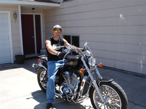1979 Ford F-150 - A rider in a black tank top, jeans, camo cap and orange-lens sunglasses sits astride a black V-twin cruiser motorcycle on a concrete drivew…