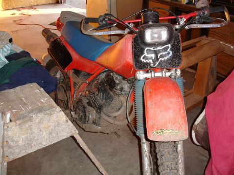 1979 Ford F-150 - Front three-quarter view of a dusty red and blue dirt bike parked in a garage, fitted with a square black rectangular headlight housing wea…