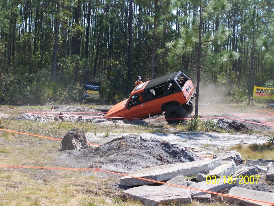 Orange off-road SUV tilted on its side in a dirt obstacle course with trees in the background. Date stamp reads 02/18/2007
