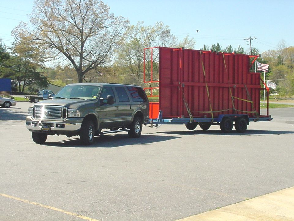 Tan Ford Excursion SUV towing a trailer loaded with red steel structures in a parking lot.