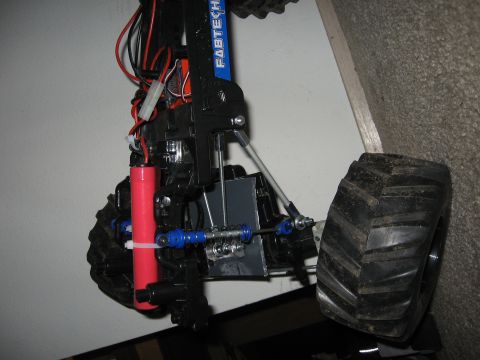 1990 Ford Bronco - Underside/side view of a bodyless RC monster truck chassis leaned against a wall, showing a red battery pack zip-tied to the black chassis…