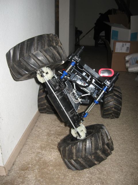 1990 Ford Bronco - Bodyless RC monster truck chassis stood on end against a wall on beige carpet, showing its dirty knobby tires, blue-anodized coil-over shoc…