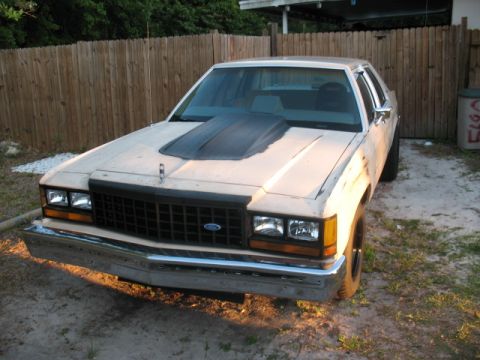 1985 Ford LTD Crown Victoria - A beige 1985 Ford Crown Victoria LTD is parked in a gravel lot with a wooden fence behind it.