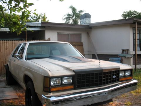 1985 Ford LTD Crown Victoria - A tan sedan with a custom hood scoop is parked in front of a house.