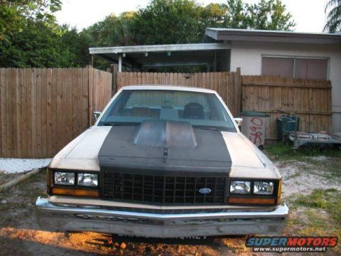 1985 Ford LTD Crown Victoria - A tan Ford Crown Victoria with a dark hood scoop is parked in front of a wooden fence and a house.