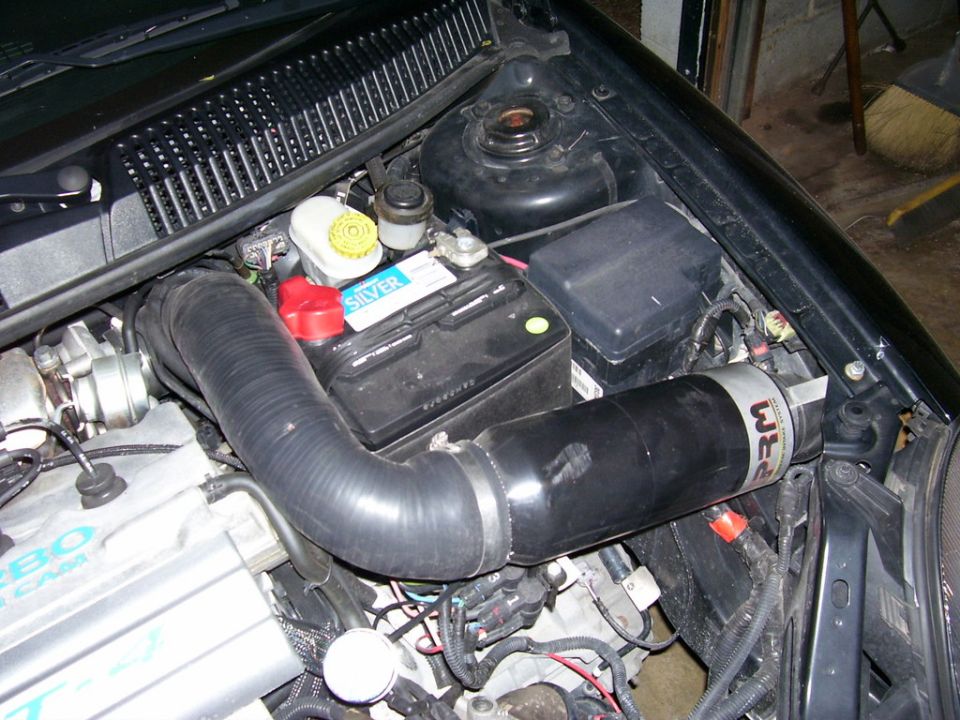 Engine bay with battery, intake tubing, and various engine components. Black hood and fender visible