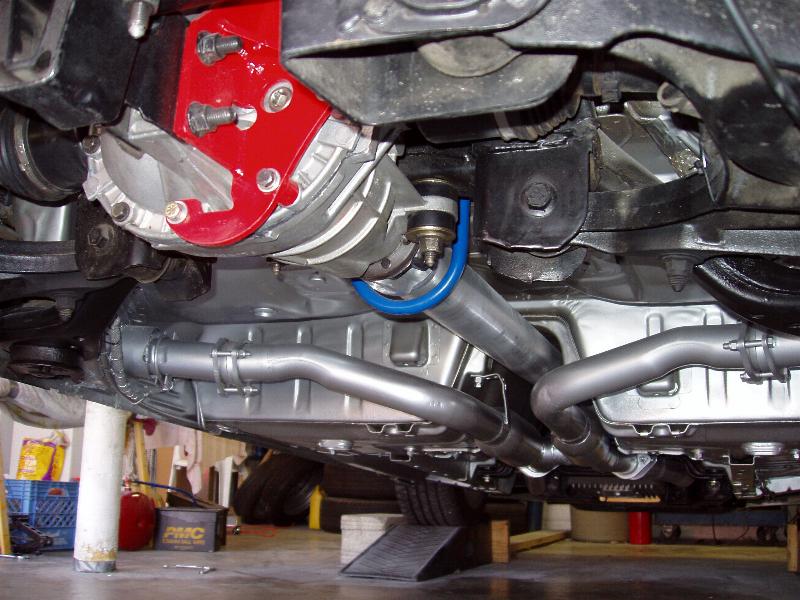 Underside of a car showing rear differential with red bracket, blue sway bar, and silver-painted exhaust pipes.