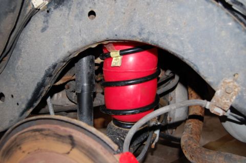 1985 Mercury Grand Marquis - Close-up of a red air spring and black coil over a shock absorber, showing rust on surrounding suspension components.