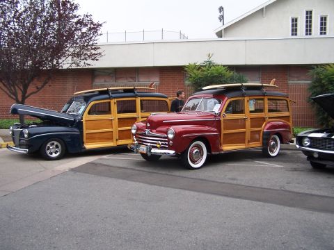 1990 Ford Bronco - Two vintage woody station wagons, one dark blue and one red, are parked next to each other with surfboards on their roofs.