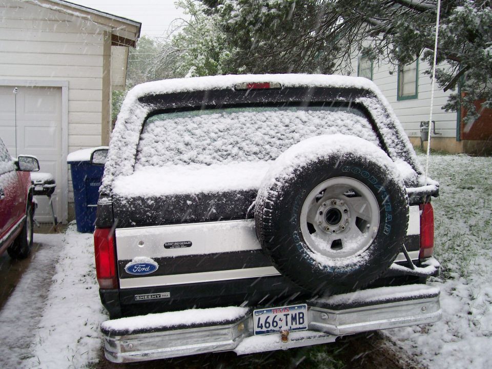 Rear of a snow-covered black Ford Bronco with spare tire and Texas plate 466-TMB in a snowy driveway