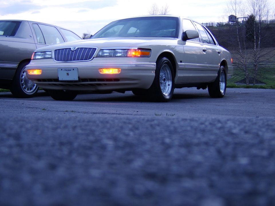 Low-angle front-3/4 view of a gold Mercury Grand Marquis sedan with lights on.