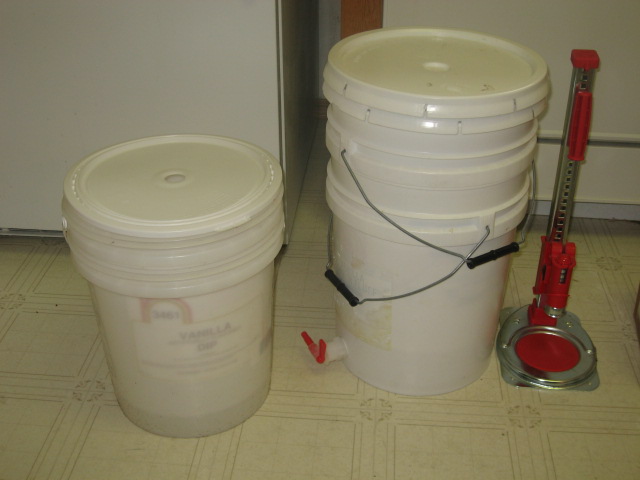 Two white 5-gallon buckets with lids on a tiled floor next to a red-handled tool.