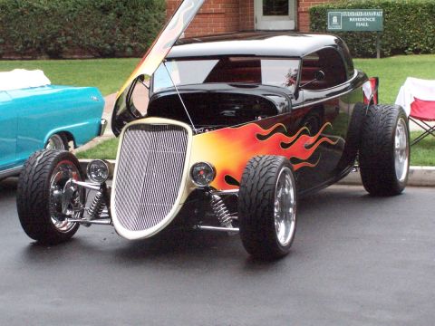 1990 Ford Bronco - A black hot rod with flame decals and its hood open sits on pavement next to a blue car.