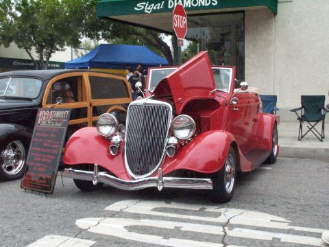 1990 Ford Bronco - A red 1934 Chevrolet convertible is displayed with its hood open at a car show, alongside a classic woodie wagon.