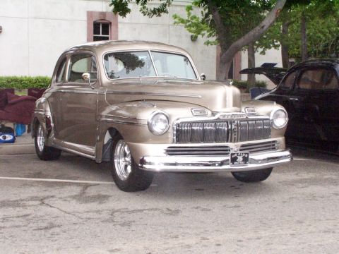 1990 Ford Bronco - A gold 1947 Mercury Coupe is parked on asphalt, with another dark car visible behind it.