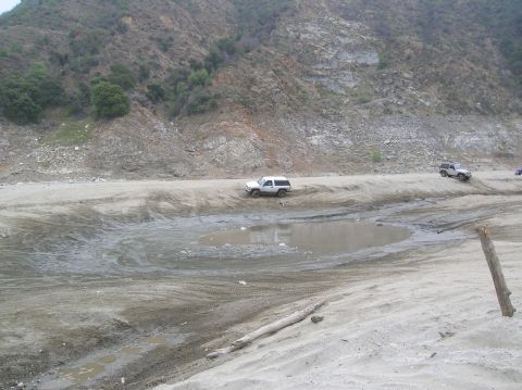 1985 Ford Bronco - Multiple SUVs navigate a challenging dirt track with muddy sections and a steep, rocky incline in the background.