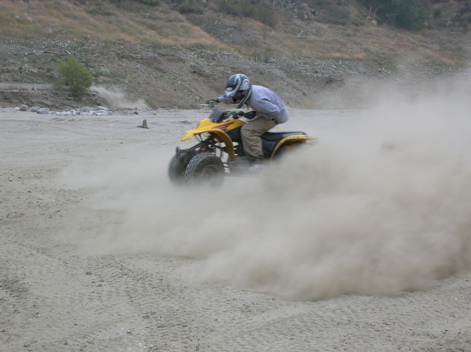 Yellow quad bike with rider wearing a helmet, kicking up a large cloud of dust on a dirt track