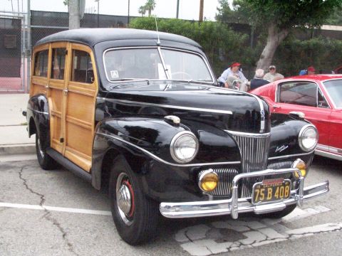 1990 Ford Bronco - A classic black woodie station wagon is parked at an outdoor event, with a red car visible to its right.