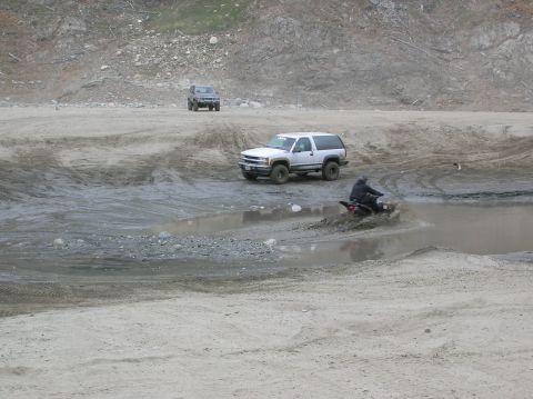 1985 Ford Bronco - Two SUVs and an ATV navigate a muddy terrain with a rocky hillside in the background.