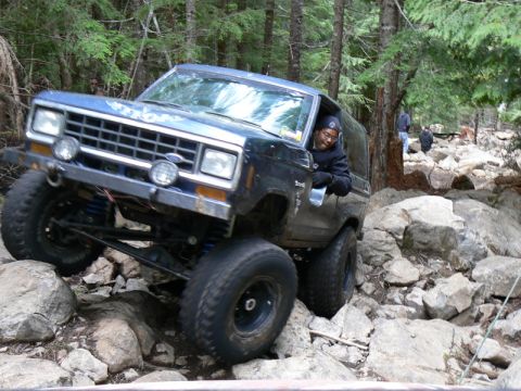 1986 Ford Bronco II - A blue Ford Bronco II is shown off-roading on a challenging rocky terrain, with trees and other vehicles in the background.