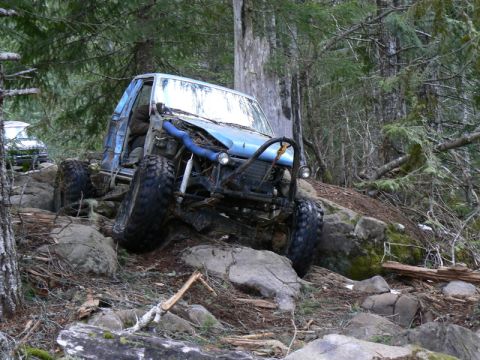 1986 Ford Bronco II - An off-road vehicle is shown navigating a challenging rocky path in a wooded area. Another vehicle is partially visible in the background.