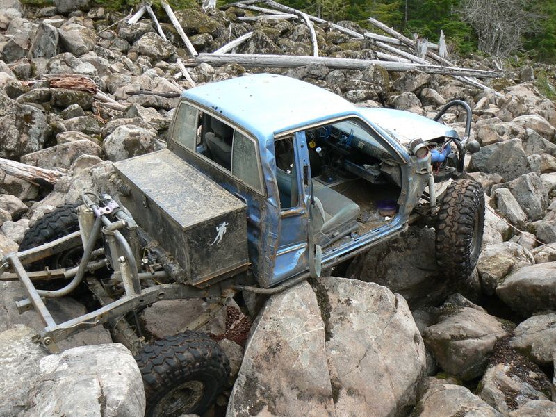 Blue off-road vehicle with its doors removed is wedged between large rocks on a wooded trail
