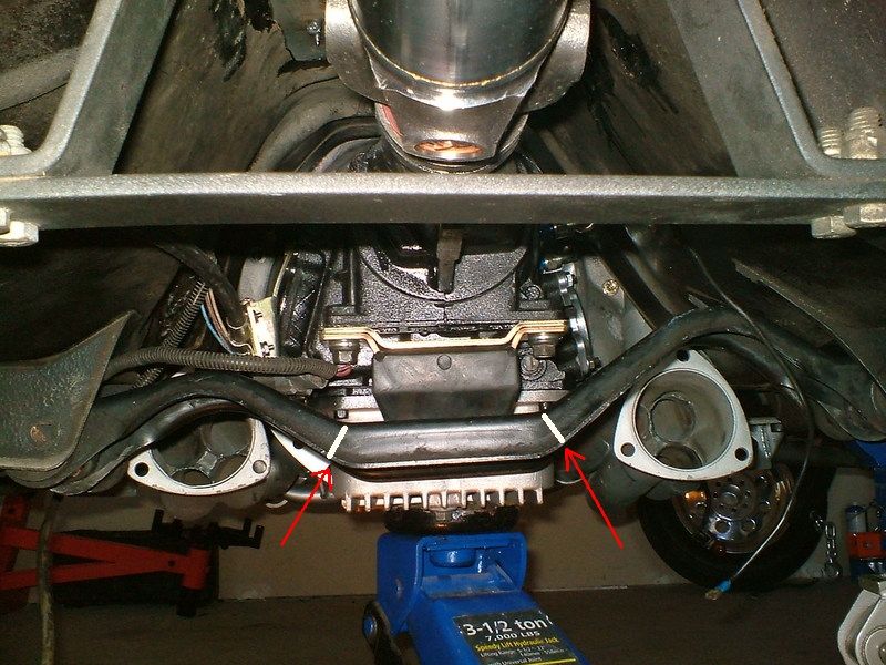 Vehicle undercarriage showing transmission, driveshaft, and exhaust pipes. A blue hydraulic jack supports the transmission