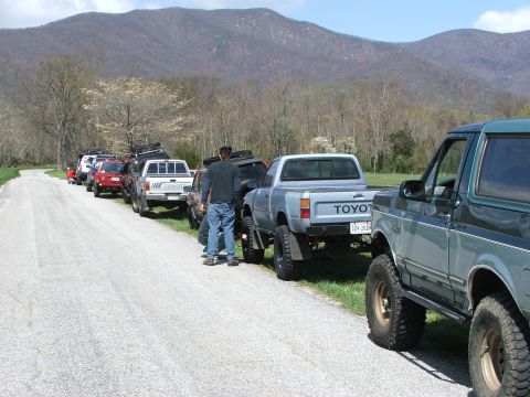 1996 Ford Bronco - Several off-road vehicles are parked in a line along a gravel road with mountains in the background.