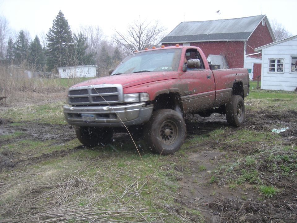 Red pickup truck covered in mud, angled front-left, with a barn and white building in the background