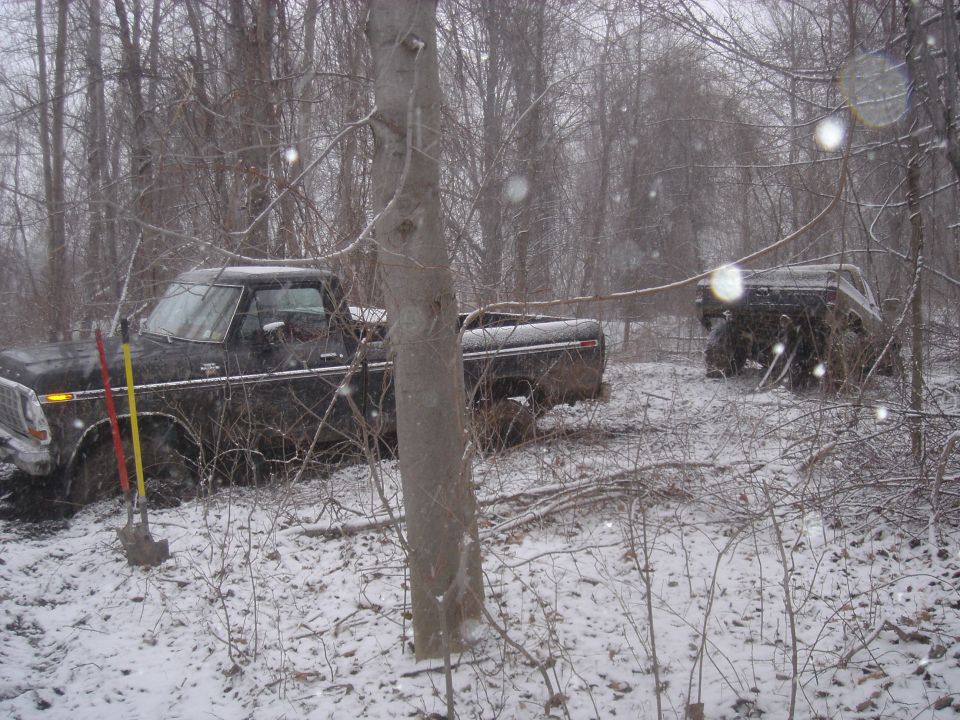 Two dark-colored trucks stuck in snowy woods; one in foreground, one in background
