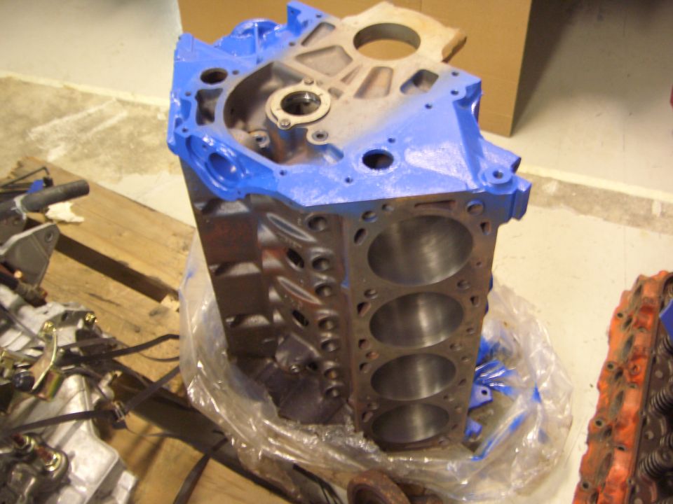 Bare V8 engine block with blue-painted end, showing cylinder bores, in a garage