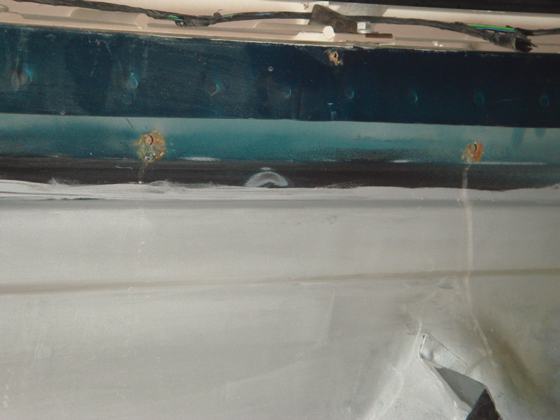 Close-up of a dark teal metal panel with rust spots and wires above. Below is a light gray surface, possibly primed or painted