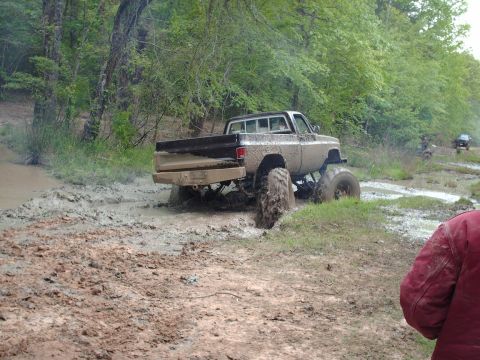1990 Ford F-150 - A lifted pickup truck navigates a challenging muddy terrain, with another vehicle visible in the background.