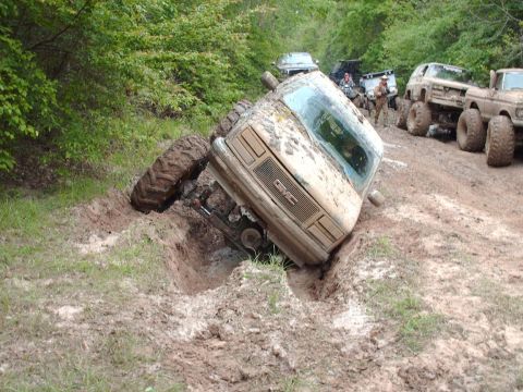 1990 Ford F-150 - Several off-road vehicles are seen in a muddy, wooded area, with one GMC truck stuck in a deep rut.