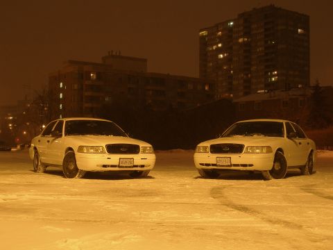 2003 Ford Crown Victoria - Two white 2003 Ford Crown Victorias are parked in a snowy lot at night with apartment buildings in the background.