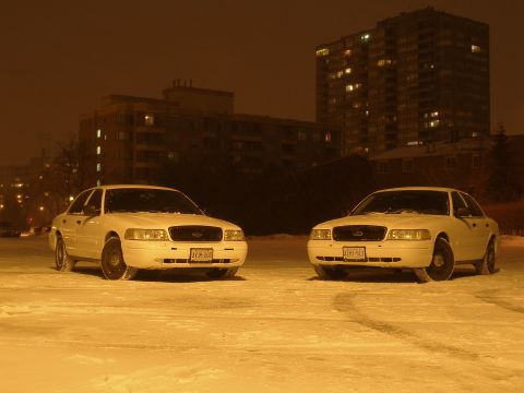 2003 Ford Crown Victoria - Two white sedans sit in a snowy lot at night with an apartment building illuminated in the background.