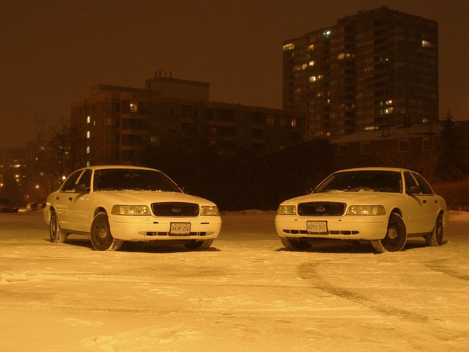 Two white sedans parked in snow at night, apartment building in background