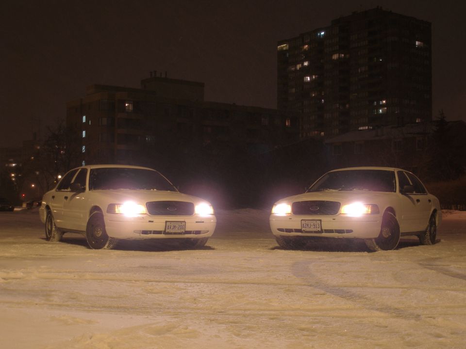 Two white 2003 Ford Crown Victorias parked in snow at night, front view