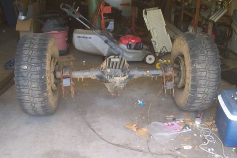 1994 Ford F-150 - Complete solid rear axle assembly with differential and mud tires sitting on a garage floor beside a lawn mower.