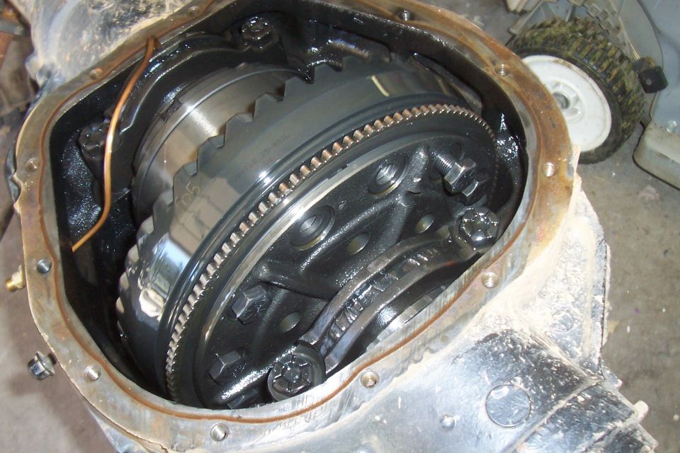 Close-up of an open differential assembly with gears and bolts visible. A lawnmower is in the background