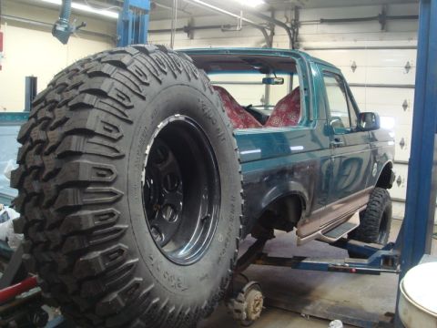 1996 Ford Bronco - Green Ford Bronco on a lift with a large spare tire, showing rust damage and missing body panels.