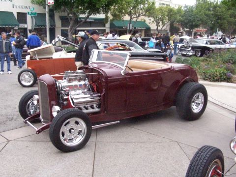 1990 Ford Bronco - A custom maroon hot rod roadster with a prominent chrome engine is displayed at a car show alongside other vehicles.
