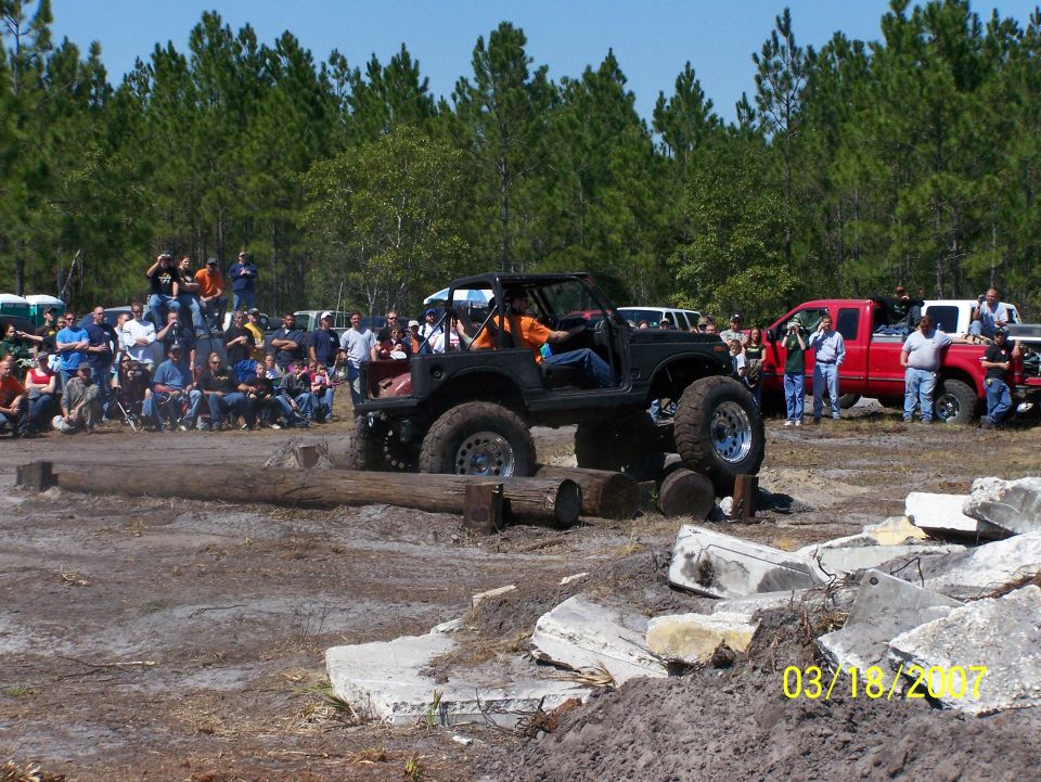 Black off-road vehicle with large tires driving over logs, crowd watching