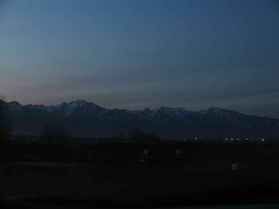Mountain range with snow-capped peaks against a dark blue twilight sky. Lights twinkle in the distance below