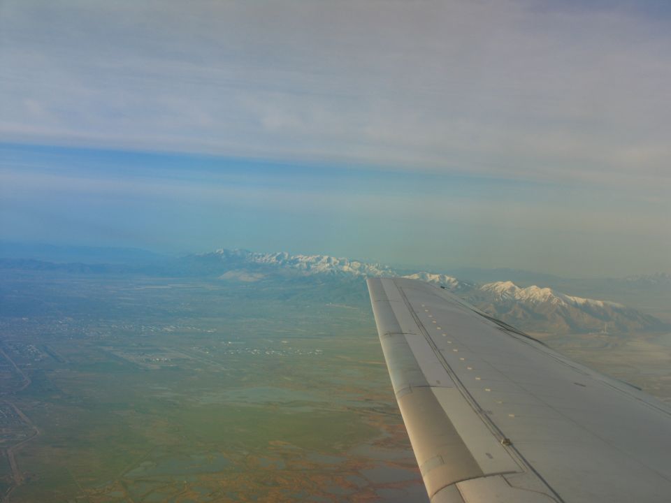 Airplane wing extends into view over a vast landscape with snow-capped mountains in the distance