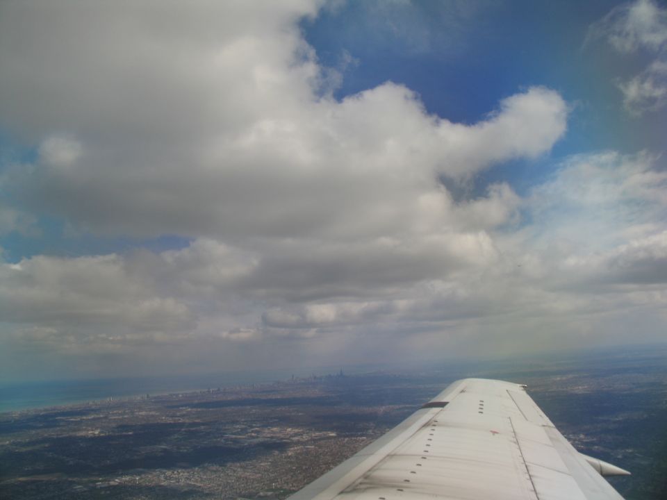 View from airplane window showing wing and clouds over a distant city