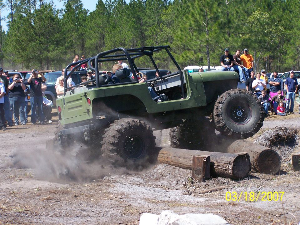 Green off-road vehicle with large tires climbing over logs, kicking up dust