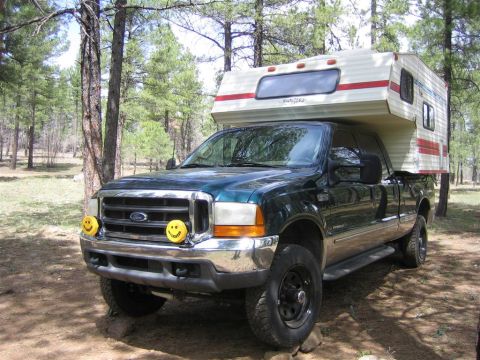 1999 Ford F-250 - A 1999 Ford F-250 Super Duty pickup truck is parked in a wooded area, fitted with a camper shell and decorative smiley face headlight cover…