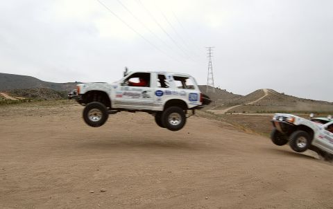 1992 Ford Explorer - Off-road trucks catch air during a race event on a dusty track with hills in the background.
