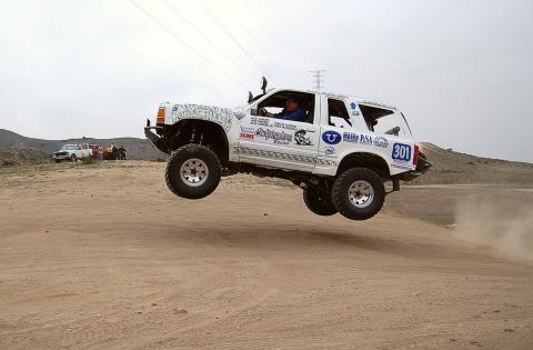 1992 Ford Explorer - Modified off-road SUV jumps over a dirt track, with other vehicles and spectators in the distance.